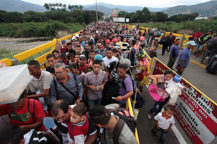 A crowd of people carrying bags walk closely, crossing a bridge, with a river and green hills behind them.