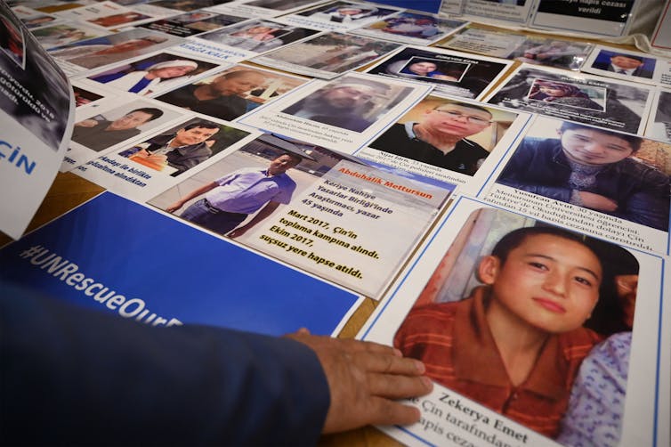 A person, whose body is out of the shot, places a hand on the photo of a young person on a table. There are many other headshots of people on the table.