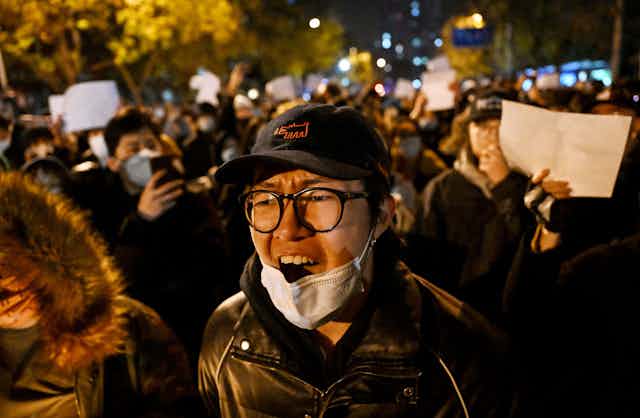A man with wearing a cap, glasses and a facemask shoots as he is surrounded by fellow protesters.