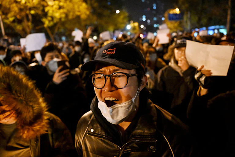 A man with wearing a cap, glasses and a facemask shoots as he is surrounded by fellow protesters.