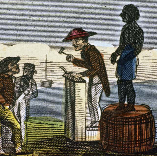 A black man stands on top of a barrel while a white man wearing a large hat with a gavel in his hand stands next to him on a podium.