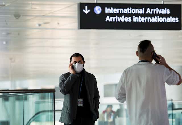 A man wearing a face mask chats on his mobile phone while standing below a sign that says 'International Arrivals' in an airport