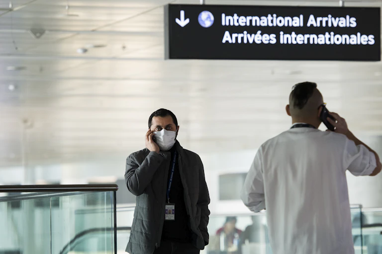A man wearing a face mask chats on his mobile phone while standing below a sign that says 'International Arrivals' in an airport