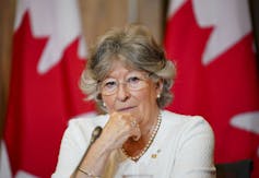 A woman with short grey-ish hair and glasses with Canadian flags behind her listens to a question at a news conference.