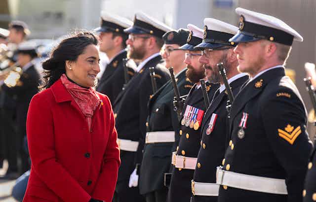 A dark-haired woman in a red coat speaks to soldiers standing in a line.