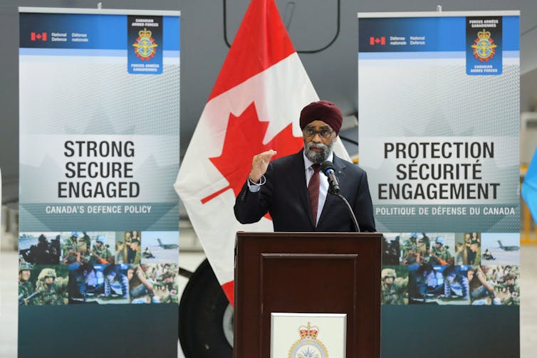 A man in a turban stands behind a podium talking into a microphone. Signs in both languages behind him read Strong, Secure, Engaged.