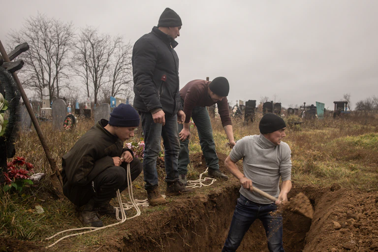 A row of four men wearing dark clothing and hats stand around and in a grave in a field on a grey day.