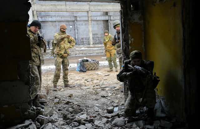 Men in camouflage uniforms stand amid rubble. One points a weapon in the direction of the camera. 