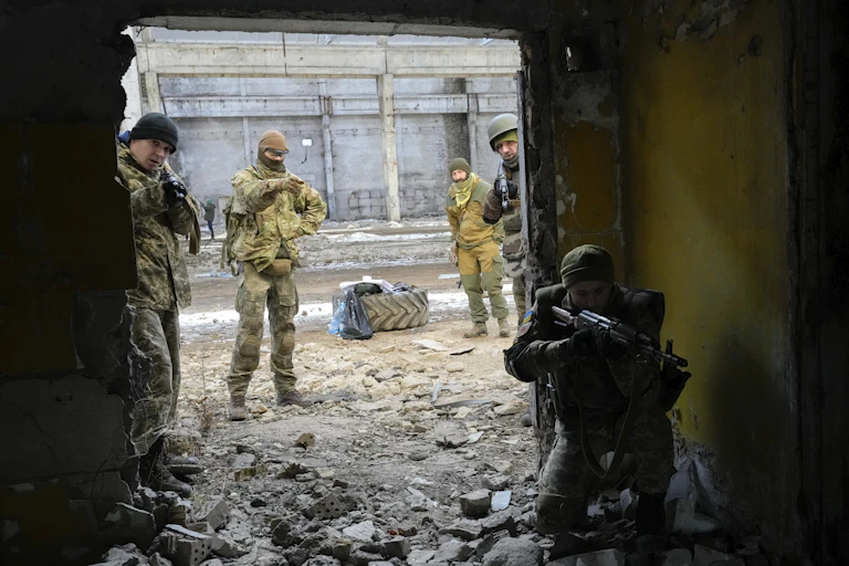 Men in camouflage uniforms stand amid rubble. One points a weapon in the direction of the camera.