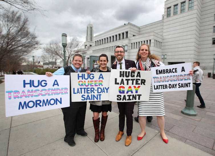 Four people holding signs stand smiling on the sidewalk outside a large white building.