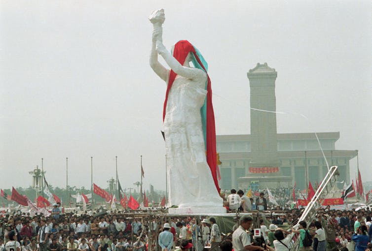 A giant white statue with arm aloft stand above 100s of people.