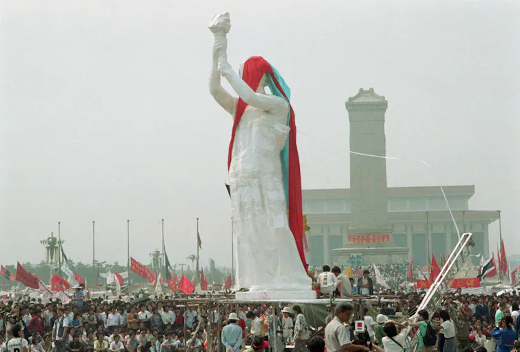 A giant white statue with arm aloft stand above 100s of people.