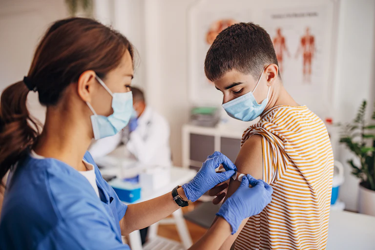 A nurse wearing a mask, scrubs and gloves places a Band Aid on a boy who has just received a COVID-19 shot.