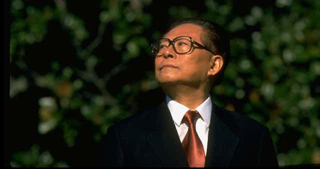 A man with crisp tie and suit and glasses looks to his right with trees behind him.