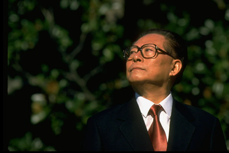 A man with crisp tie and suit and glasses looks to his right with trees behind him.