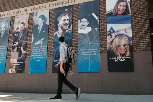 A man wearing a backpack walks down a sidewalk next to a brick wall with big posters advertising a university.