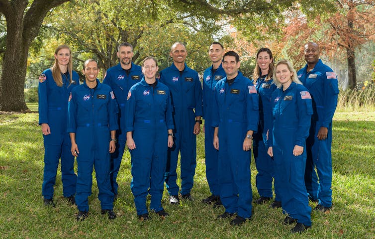 A group of ten diverse people in dark blue jumpsuits standing outdoors on a sunny day