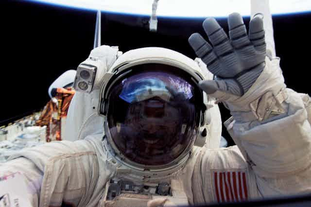 An astronaut in a white suite waving with a rubber gloved hand, face invisible behind a reflective dark visor