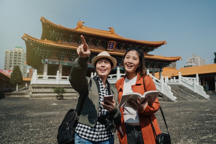 Two young women tourists holding guidebooks, cameras and cell phones, pointing and smiling with an east Asian temple behind them