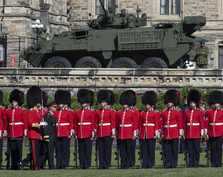 A line of soldiers in red serge and black fur hats stand in a line, a large armed vehicle behind them.