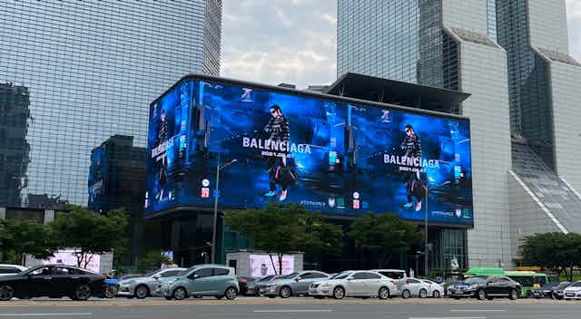 A bright Balenciaga billboard in the middle of a busy road. Model wears all black against a blue background.