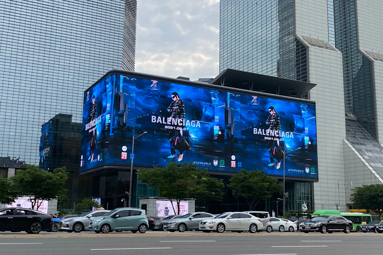 A bright Balenciaga billboard in the middle of a busy road. Model wears all black against a blue background.