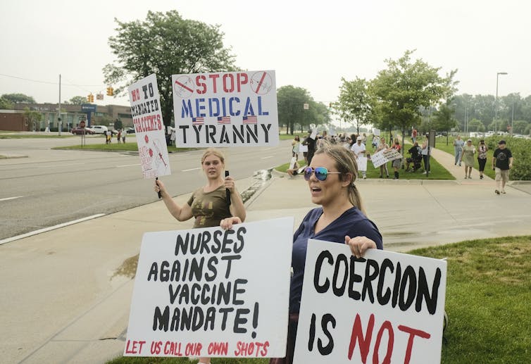 Two nurse protesters stand near a street holding up signs arguing against vaccine mandates, with a large group of protesters in the background.