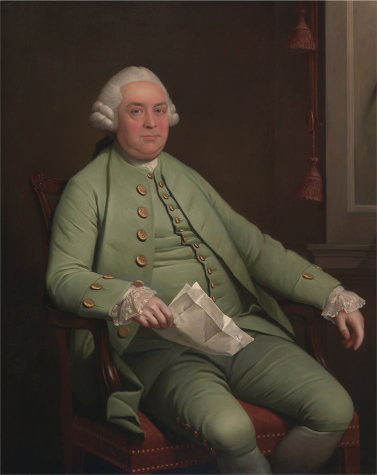 A man with a white wig and dressed in an 18th century light green suit, sitting with a piece of paper in his hand.