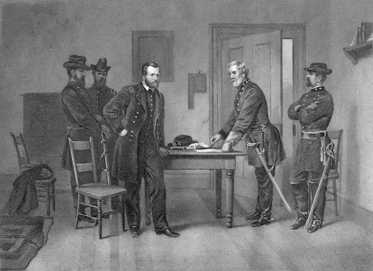 A white man dressed in a dark military uniform stands at a table with another white man dressed in a military uniform.