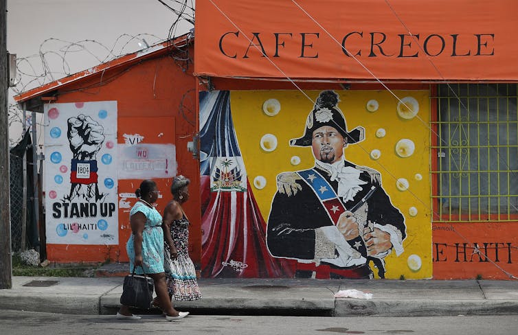 Two women walk past Cafe Creole, with vibrant paintings on the side, including one wall reading 'Stand up lil Haiti' with a raised fist.