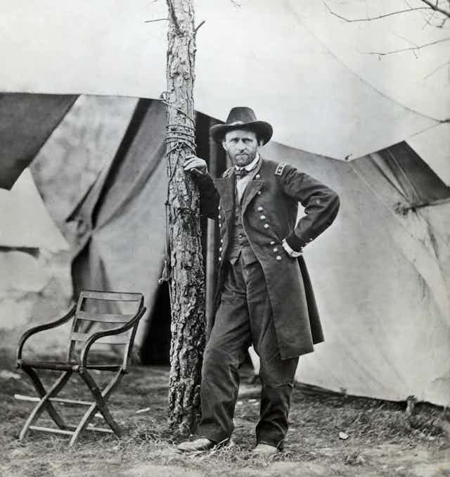 A white man wearing a dark military uniform leans against a tree near some tents. 