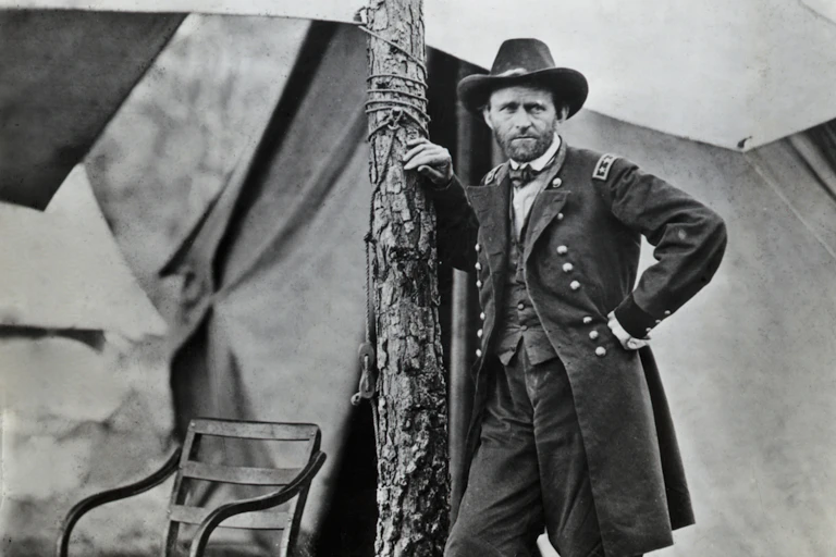 A white man wearing a dark military uniform leans against a tree near some tents.