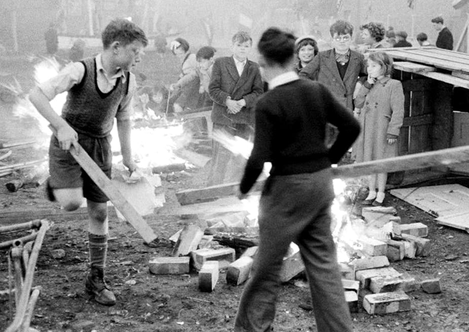 Black and white shot of boys in white shirts and vests putting wood on a bonfire.