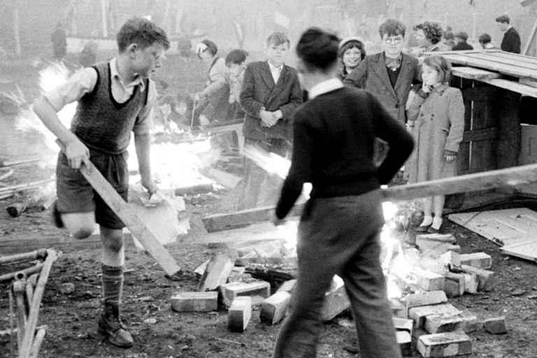 Black and white shot of boys in white shirts and vests putting wood on a bonfire.