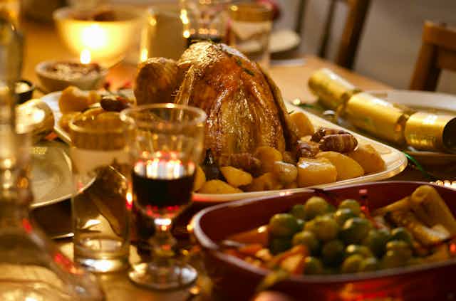 A table laden with various cutlery, drinks and meals, including roast chicken and potatoes, a bowl of peas and a glass of wine. 