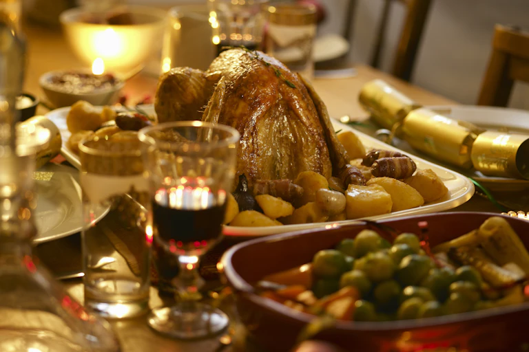 A table laden with various cutlery, drinks and meals, including roast chicken and potatoes, a bowl of peas and a glass of wine.