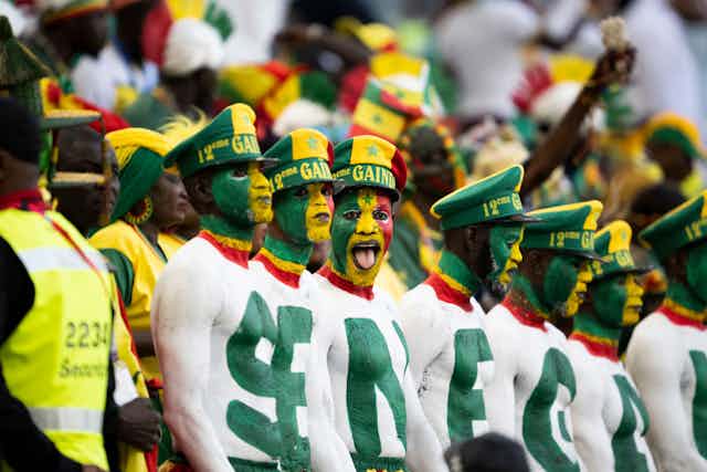 Fans in a stadium, a row of men with their upper bodies painted in green and white, each bearing a letter to spell Senegal. 