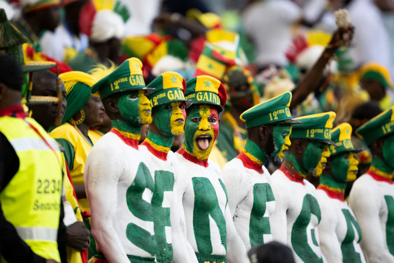 Fans in a stadium, a row of men with their upper bodies painted in green and white, each bearing a letter to spell Senegal.
