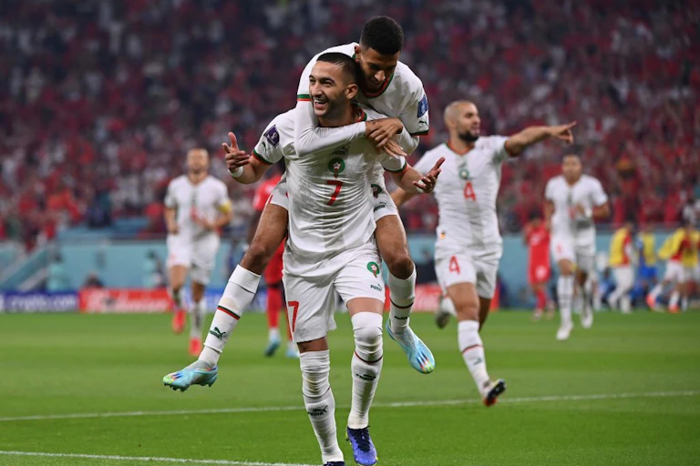 Football players in the colours of Morocco celebrate. One player jumps onto the shoulders of another as they run forward.