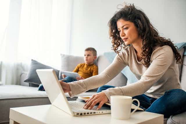 Une femme, assise sur un sofa, travaille sur son ordinateur, un jeune enfant à ses côtés