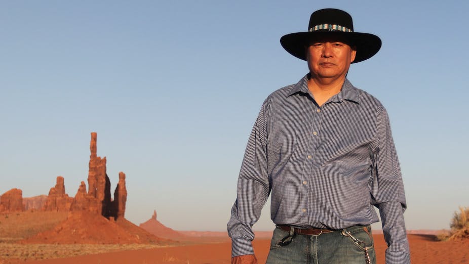 Standing against a pale blue sky and the rust-colored ground and rock formations of the desert, a Native American in black hat, grey shirt and blue jeans poses for the camera.