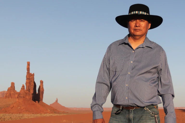 Standing against a pale blue sky and the rust-colored ground and rock formations of the desert, a Native American in black hat, grey shirt and blue jeans poses for the camera.
