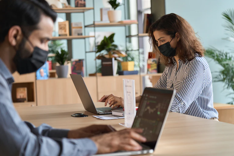 Two people sit at a dining table with their laptops and they both well face masks.
