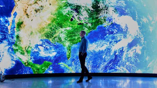 A man walks in front a giant screen showing North America from space.