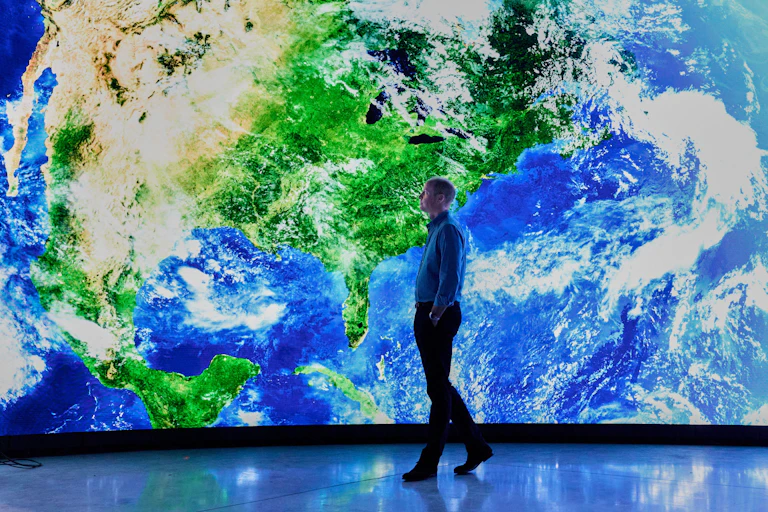 A man walks in front a giant screen showing North America from space.