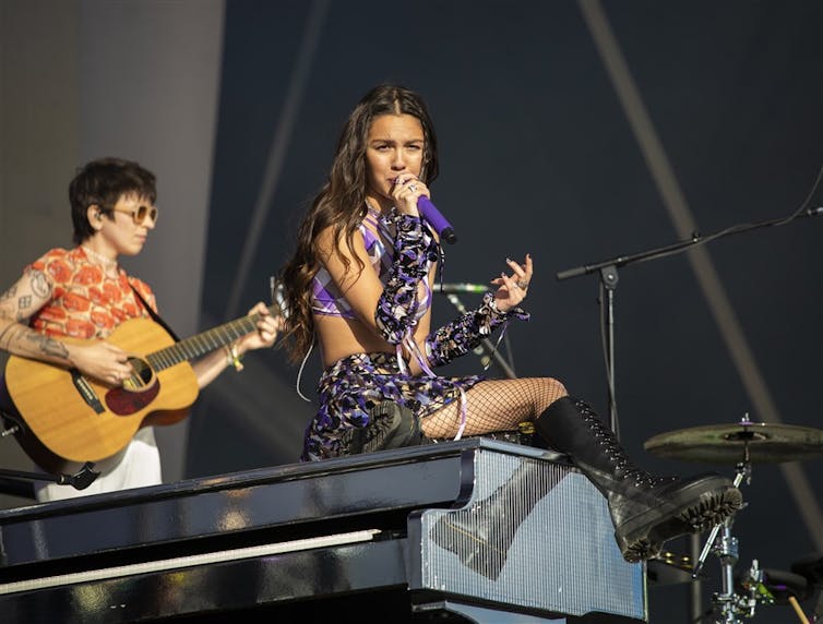 Olivia Rodrigo sits on a piano holding a microphone during her 2022 Glastonbury set. She wears a plaid two piece and fishnet tights.
