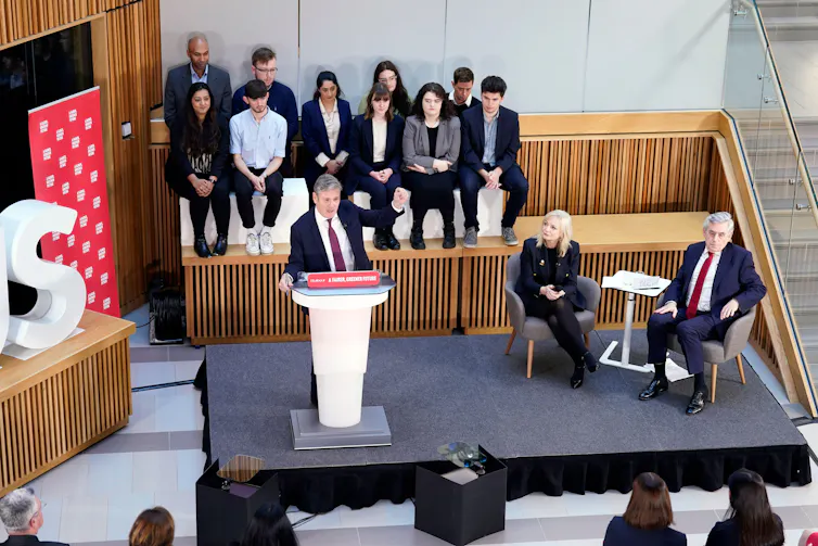 Gordon Brown giving a speech while Tracy Brabin and Keir Starmer sit at a table on the same stage.