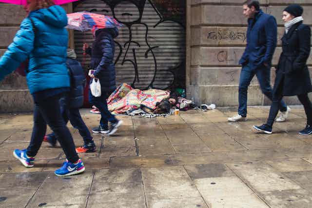 Pedestrians pass in front of a person sleeping in the street covered with blankets.