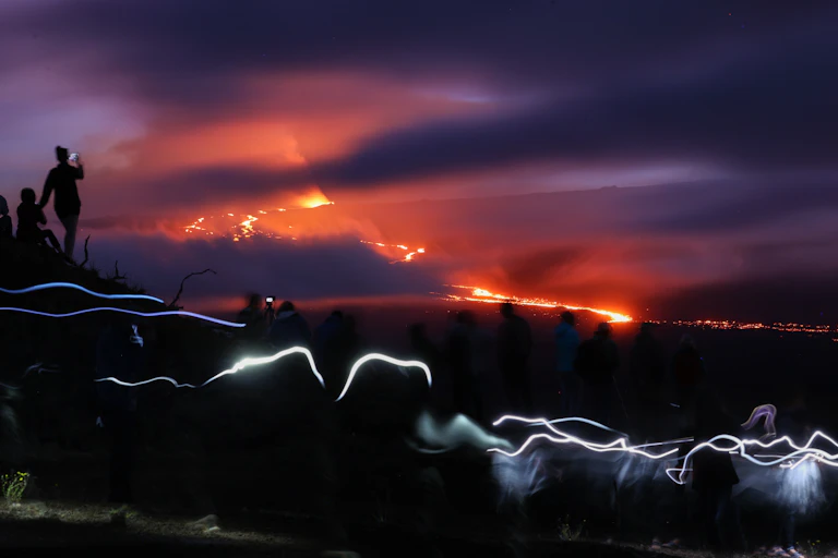People observing the fire and lava flowing from an erupting volcano, and the sky is full of red, purple and orange colors.