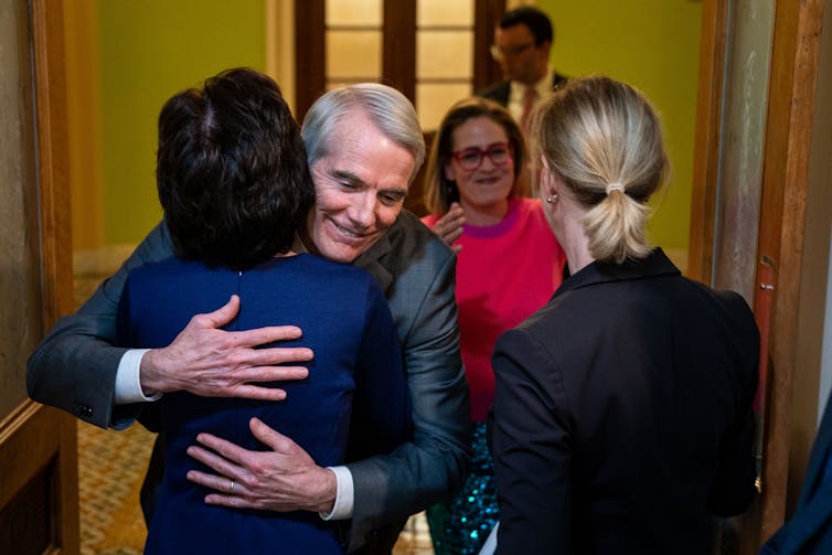 A white man in a suit is seen hugging a lady with dark hair. Next to him a blond woman with red glasses moves to hug another woman with blonde hair, as seen from the back.